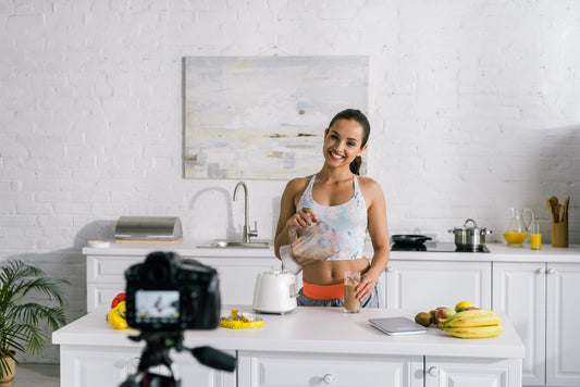 A fitness influencer recording in their kitchen, pouring a smoothie into a glass and smiling at the camera.